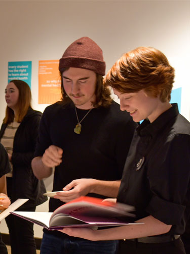 Students stand in an art gallery with posters on the walls, and look over design books