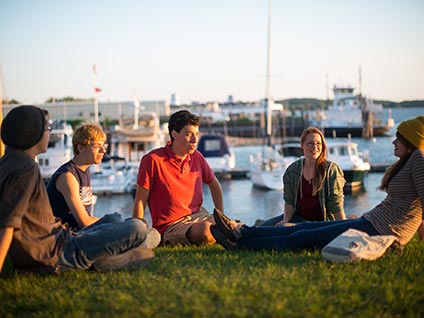 Students from Champlain College in Burlington, Vermont, sitting on the grass with sailboats in the background on the shore of Lake Champlain.
