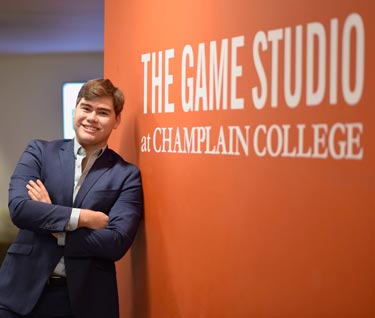 Student poses in front of a wall on which is written "The Game Studio at Champlain College"