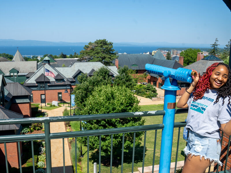 Student looking and laughing over her shoulder at an offscreen speaker after looking out at Lake Champlain through telescope on the library terrace