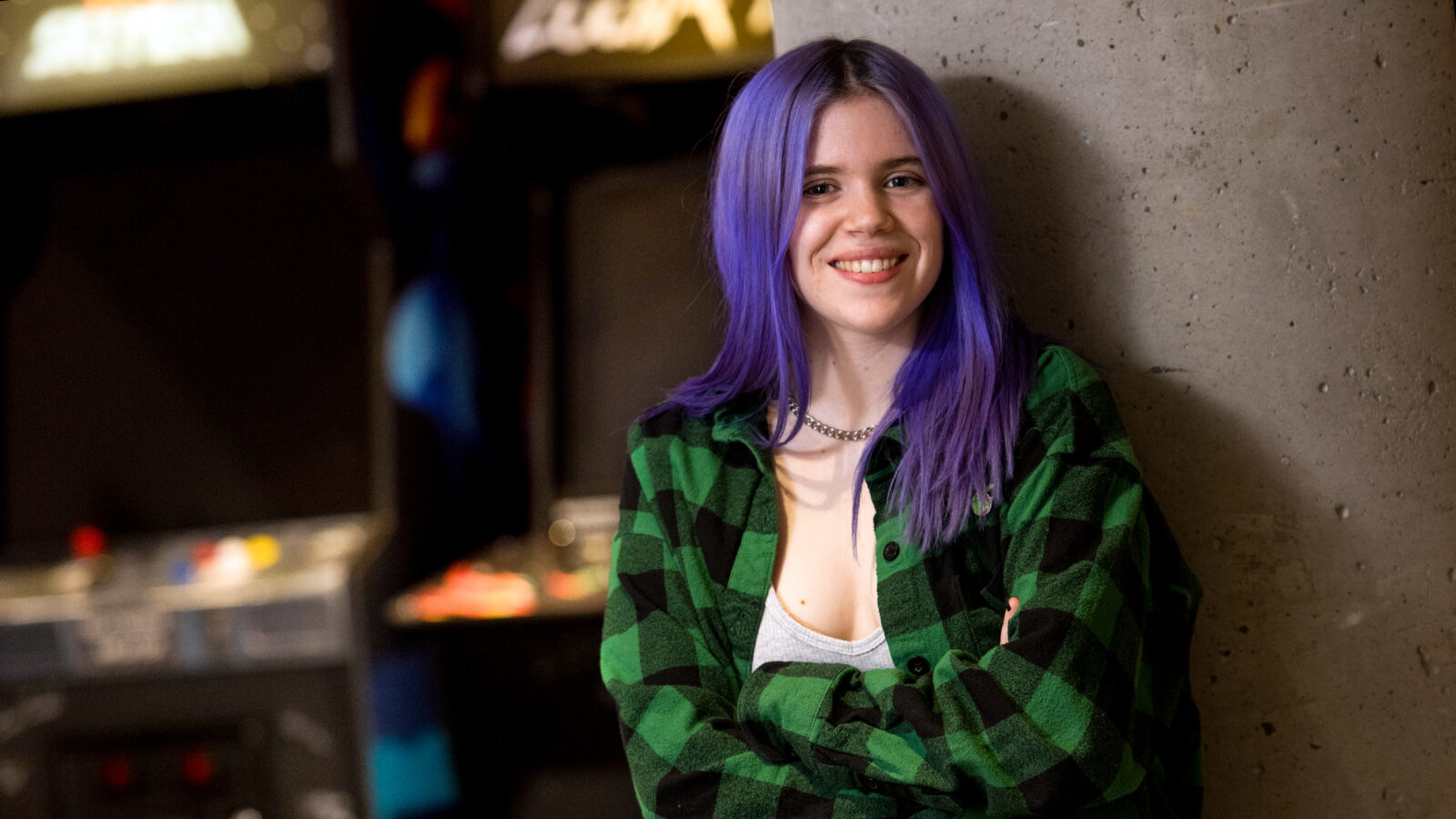 female student with purple hair stands in front of arcade games, smile at camera