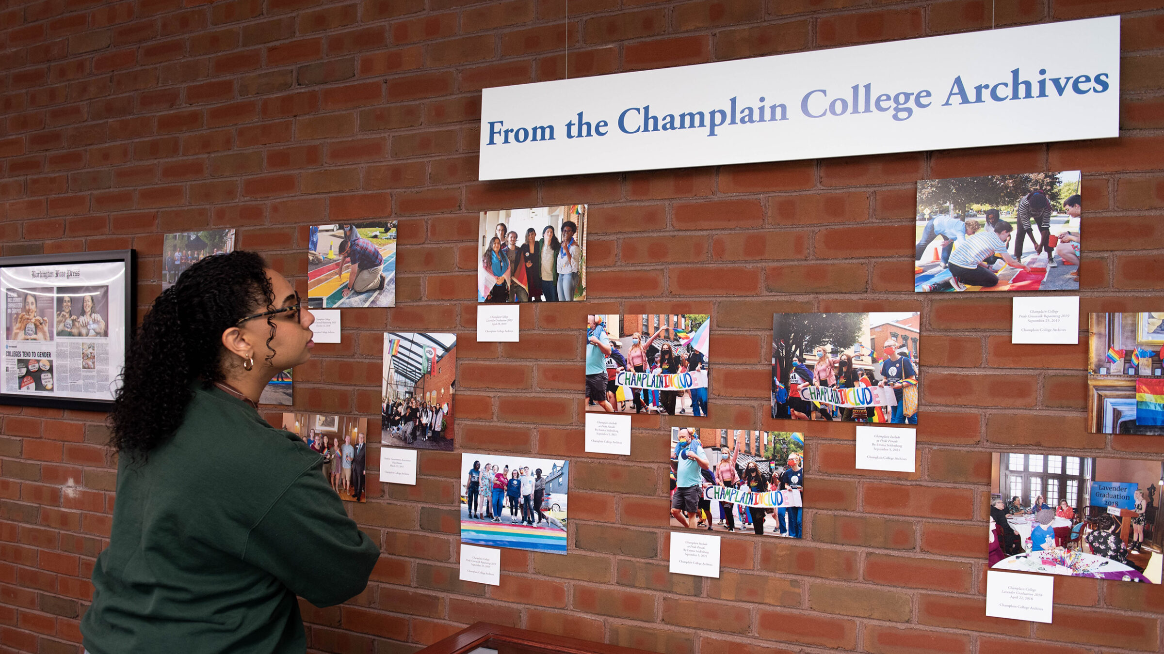 a student looks at photos from the Champlain College Archives hung on a brick wall. The photographs depict contemporary people and are quite colorful with many rainbows.