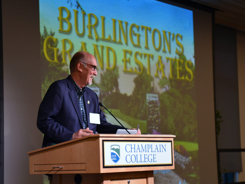 A speaker stands at a Champlain College podium. A presentation titled Burlington's Grand Estates is on a large screen behind him.