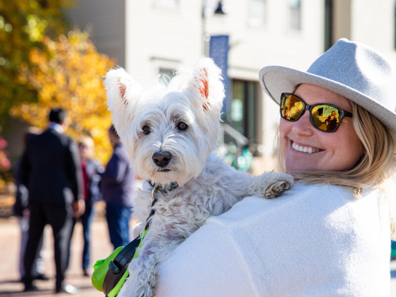 woman in a hat and sunglasses holds her dog on her shoulder