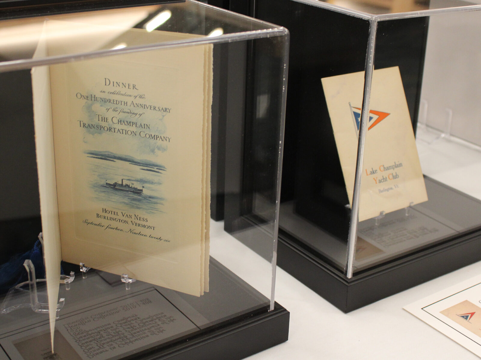 Menu for the 100th anniversary of the Champlain Transportation Company and pamphlet for the Lake Champlain Yacht club on stands under display glass.