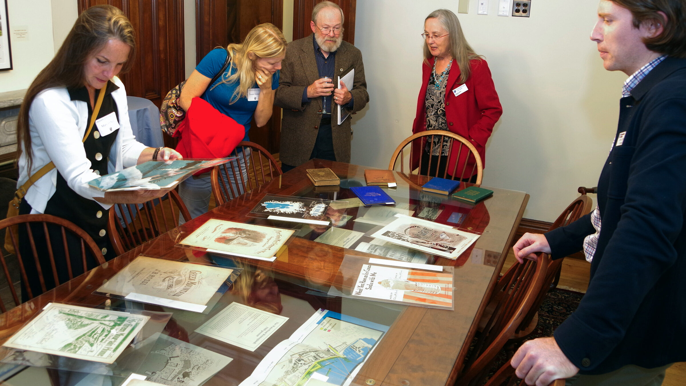 Several people stand around a table looking at sheet music covers, maps, and books on display both on top of the table and under a glass tabletop.