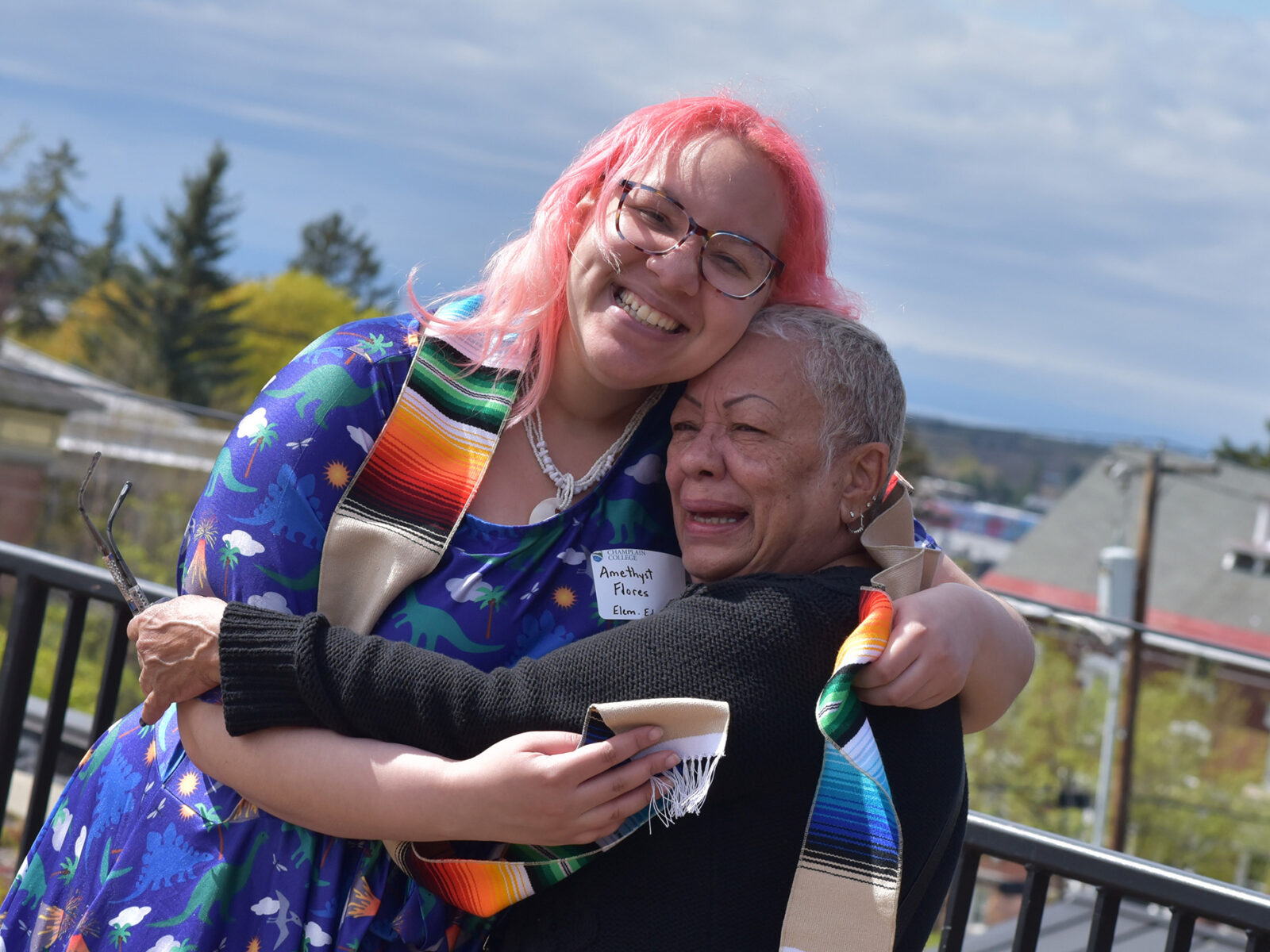 student with pink hair and commencement stole hugs other family member, all smiles