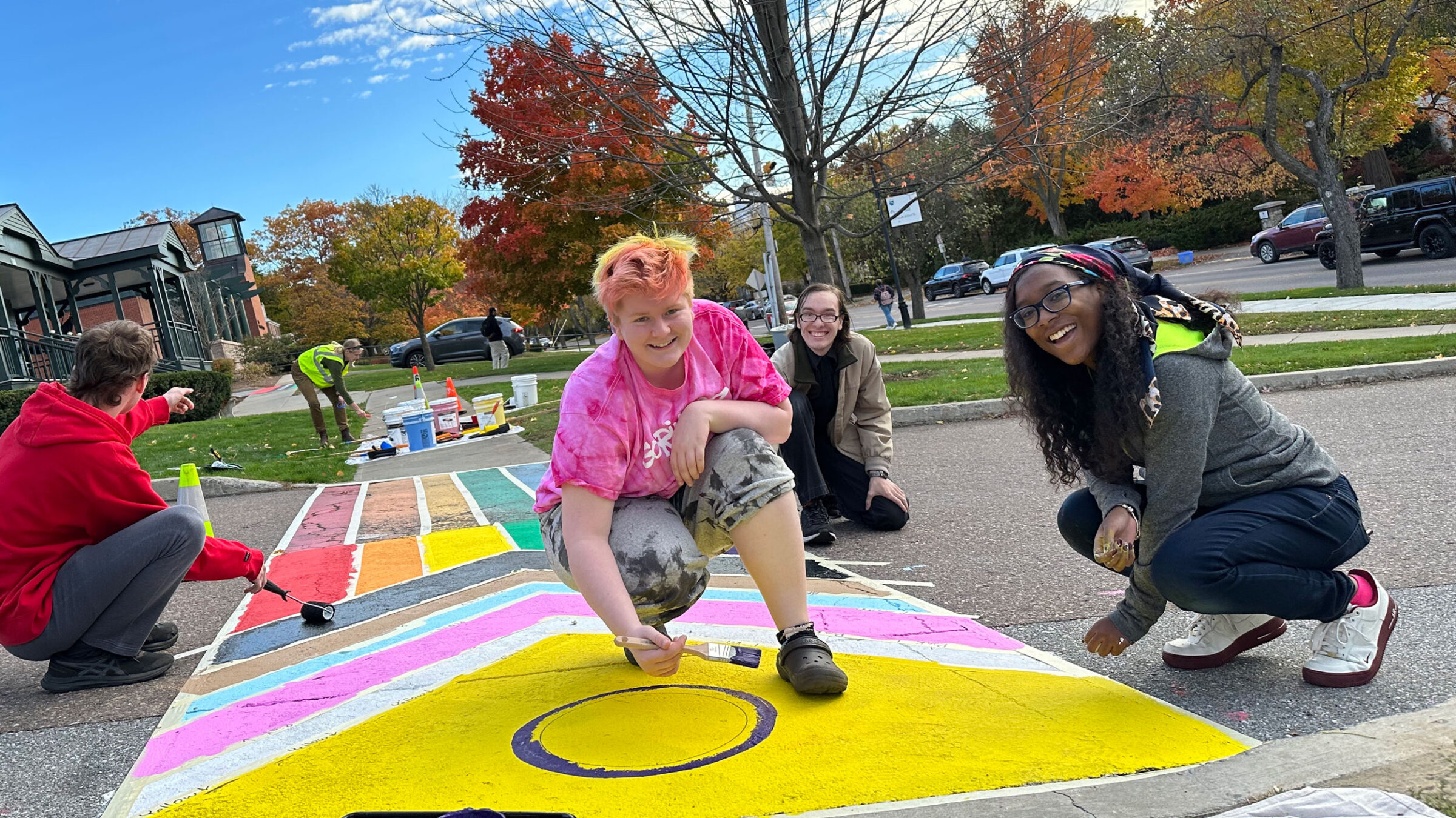 four students smile with paintbrushes in hand as they paint an intersex flag crosswalk on campus, with blue skies behind them