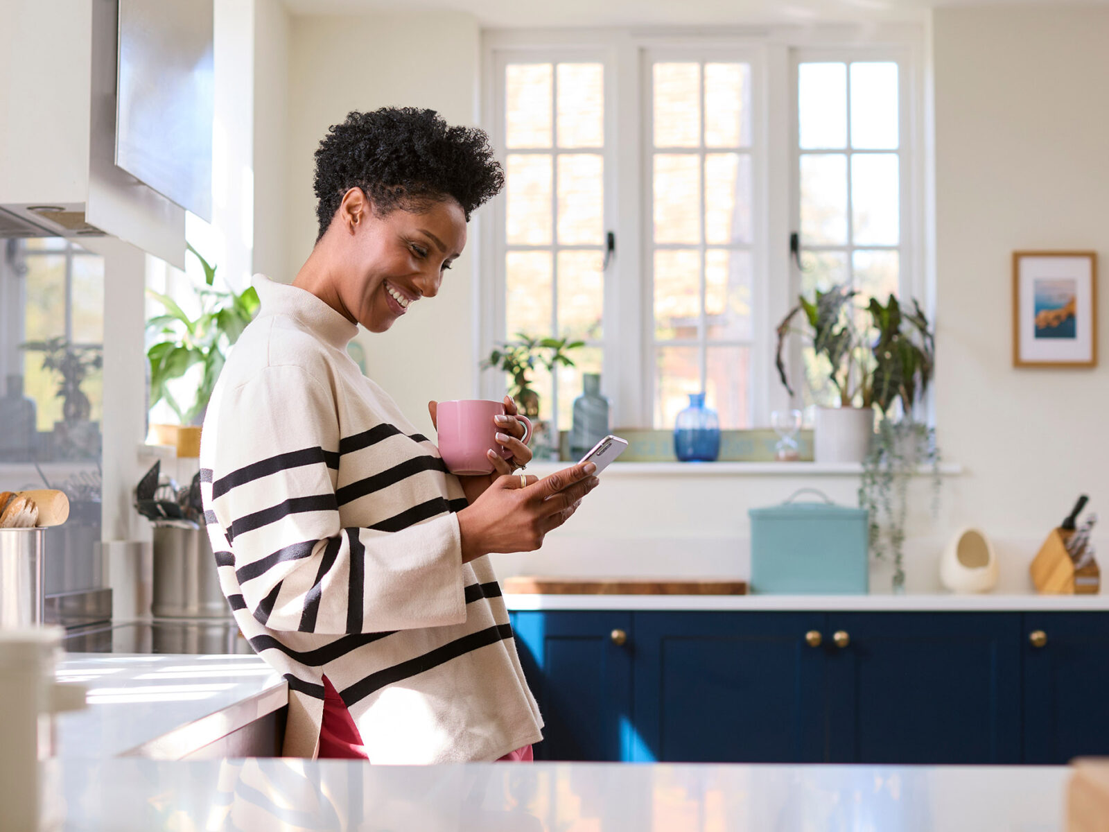 Mature Woman At Home In Kitchen Drinking Coffee And Checking Social Media On Mobile Phone
