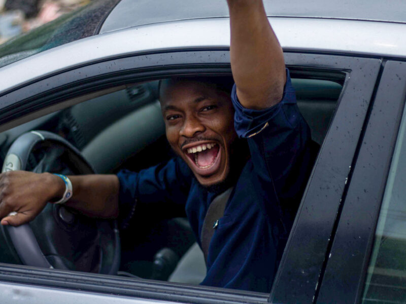 individual waving at the camera from their car window with a large grin