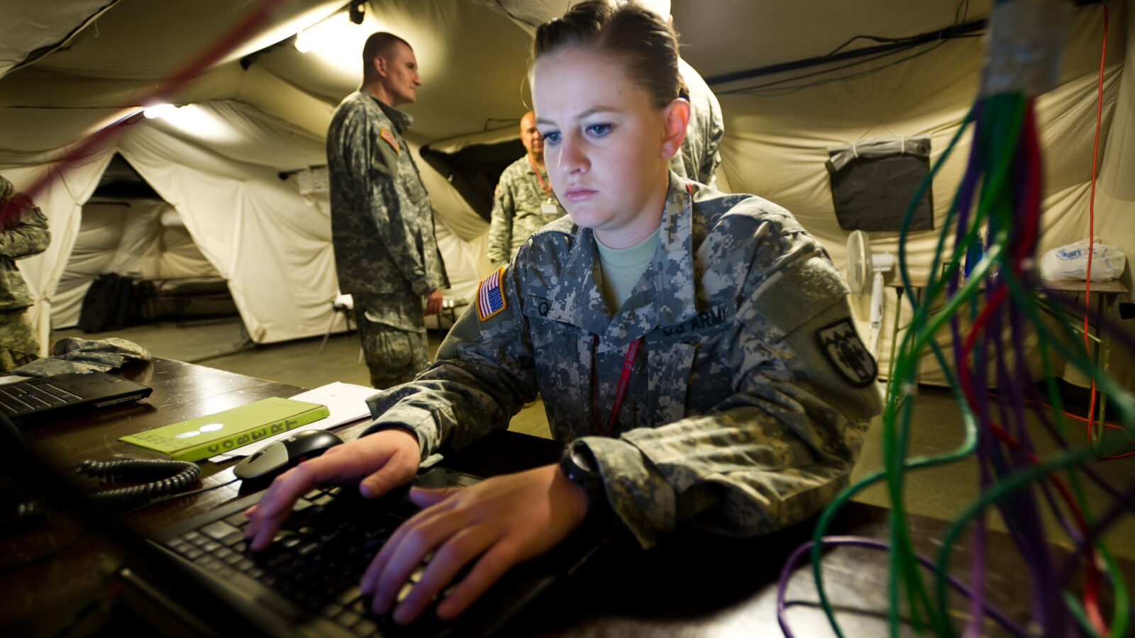 a military person uses a laptop in a tent, commanders in background