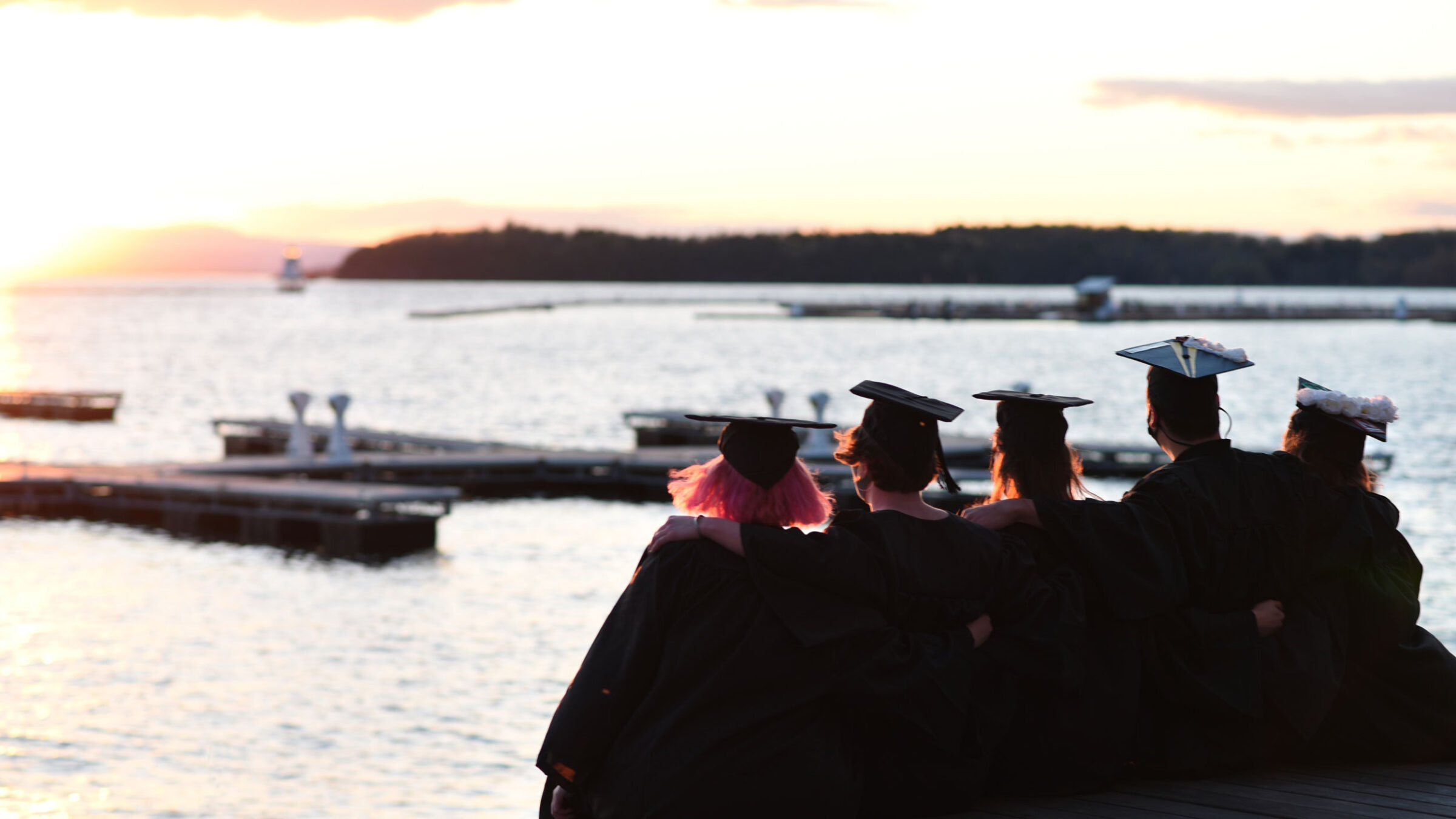Five Champlain graduates huddle together and watch the sunset over Lake Champlain in their caps and gowns.