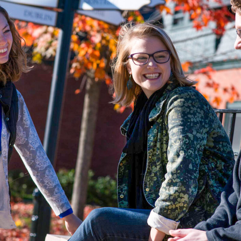 a group of students sitting outside in the fall smiling at each other