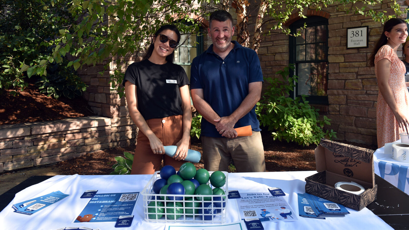 two champlain college counseling staff members stand and smile behind a table at the fall activities fair. a box of stress balls sit on the table covered in a white tablecloth.