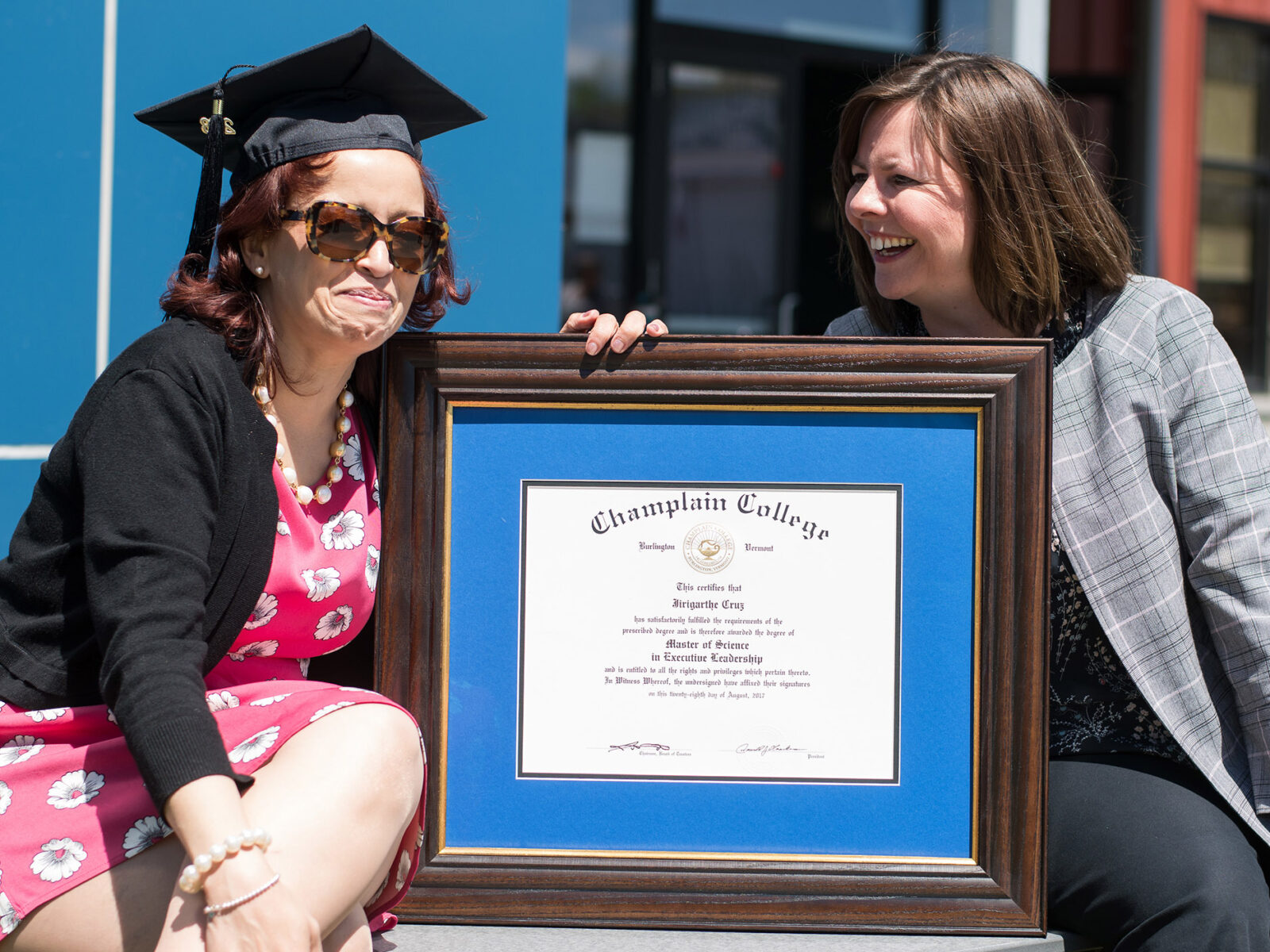 an adult graduate and another woman pose with a framed diploma