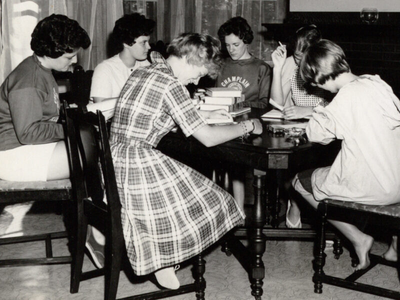 Black and white photograph depicting a group of Champlain College students studying in the common room of Jensen Hall. Six female students are seated at a table, bent over their textbooks and notes.