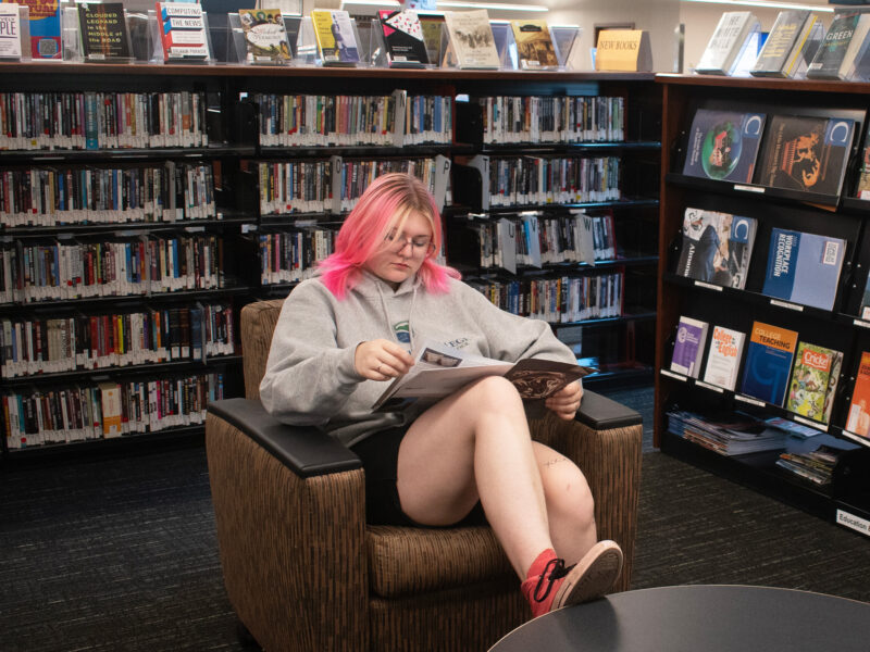 Student sitting down reading a magazine with shelves of magazines and DVDs in the background.
