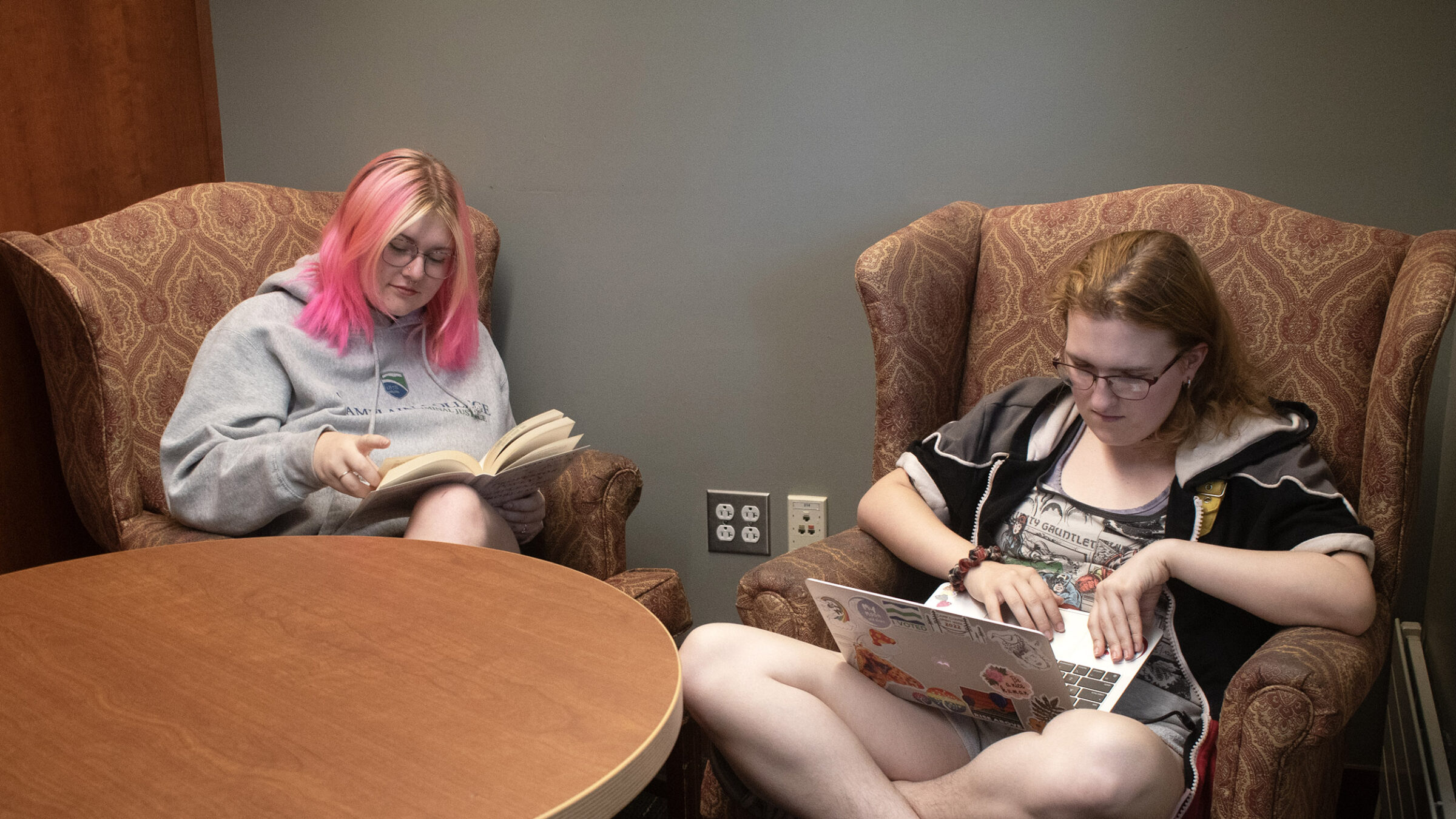 Two students sitting in comfortable armchairs in the library. One is reading while the other is on a laptop.