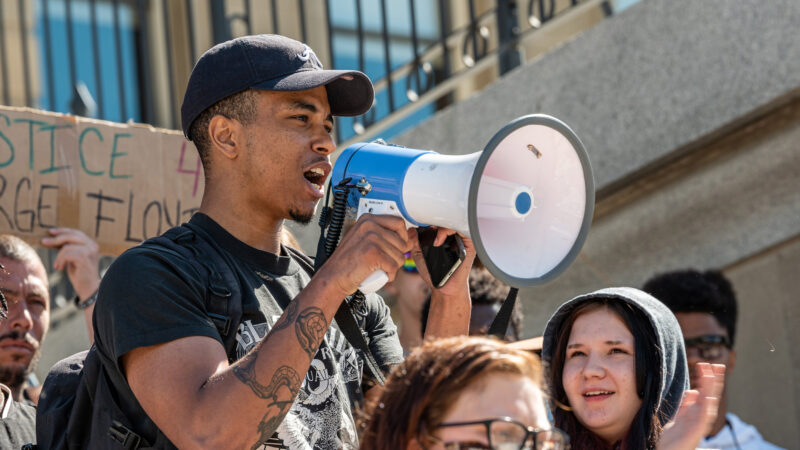 a crowd gathers on the stairs of a building; man with a bullhorn