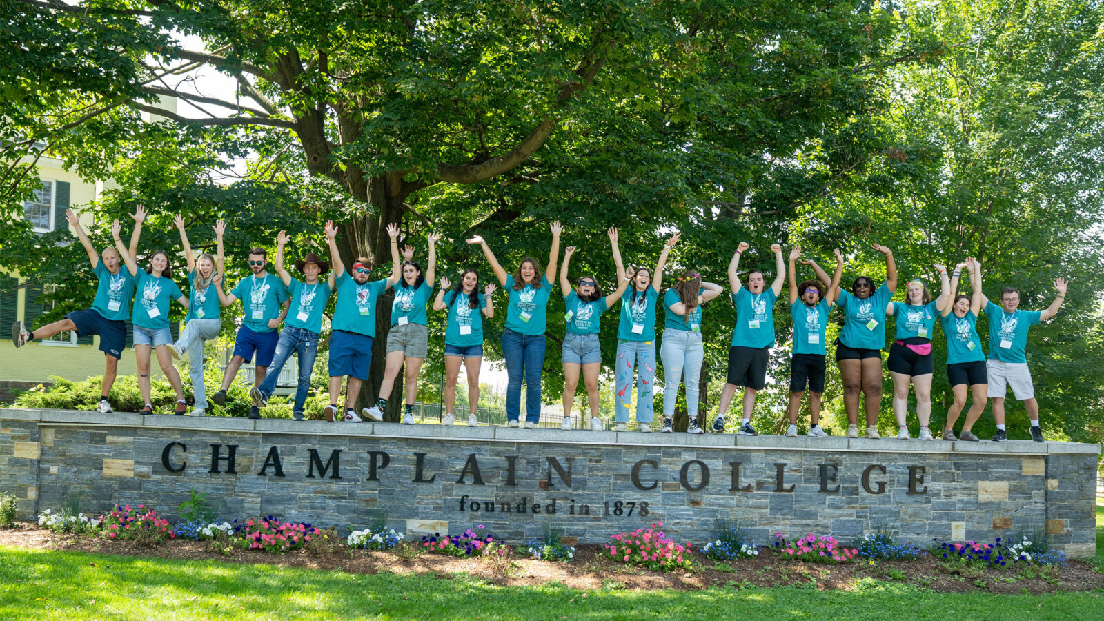 a group of 19 orientation leaders wearing matching blue shirts stand on a short wall that says "champlain college," raising their hands in the air and cheering
