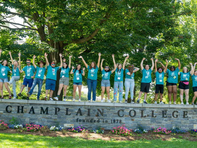 a group of 19 orientation leaders wearing matching blue shirts stand on a short wall that says "champlain college," raising their hands in the air and cheering