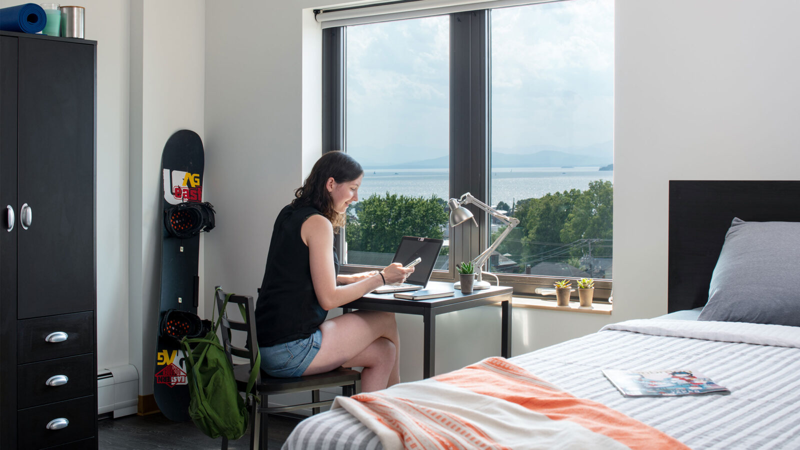 student sits at a desk in 194 residence hall room. the desk is next to a single bed and a lake view is out the desk window