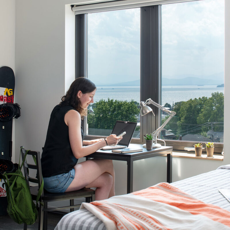 student sits at a desk in 194 residence hall room. the desk is next to a single bed and a lake view is out the desk window