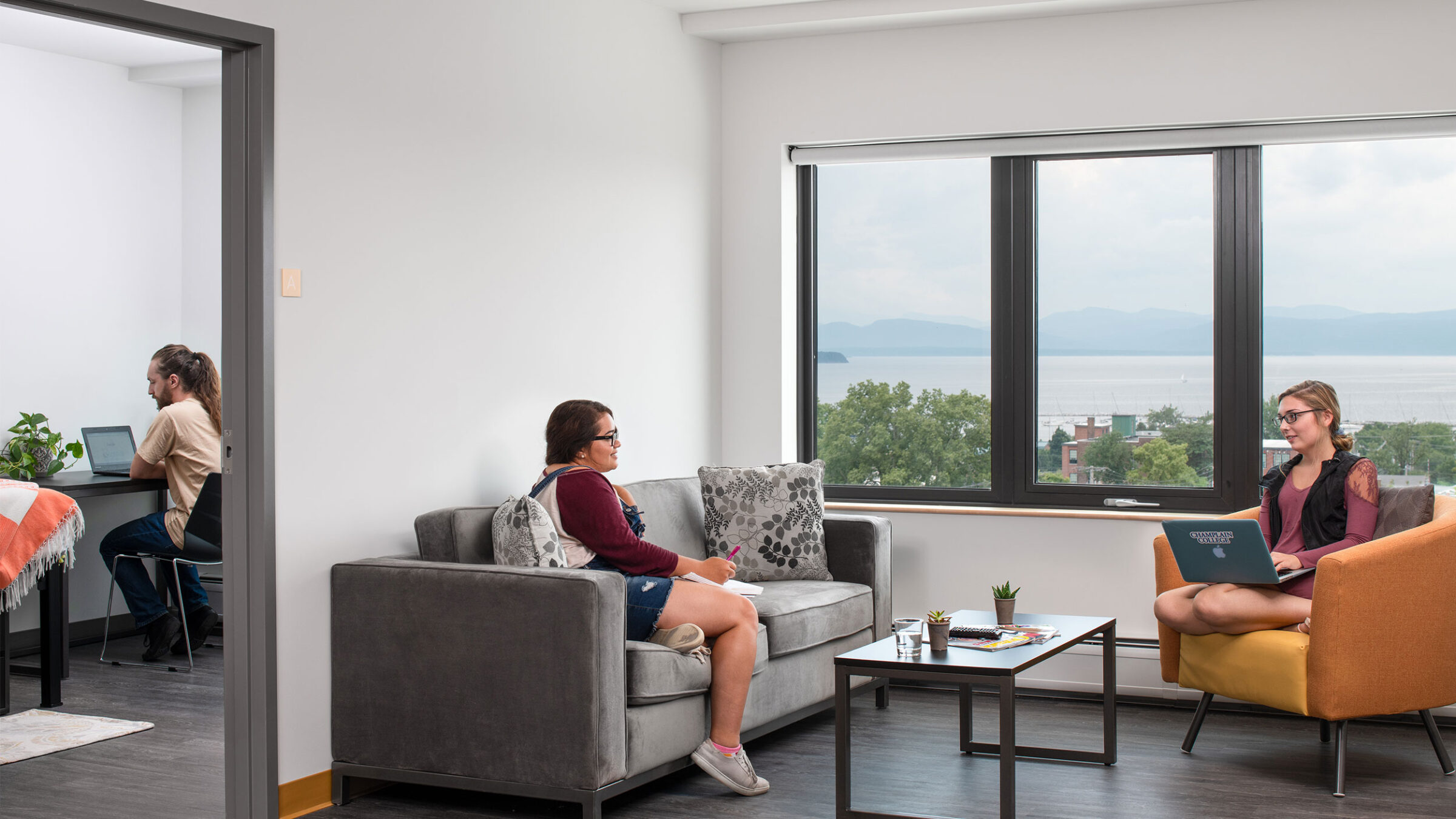 two students sit and chat in a 194 common room overlooking lake champlain while a third students sits at a desk in the bedroom on a laptop