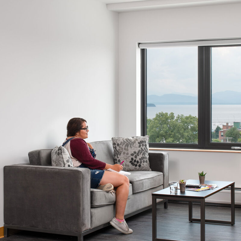 two students sit and chat in a 194 common room overlooking lake champlain while a third students sits at a desk in the bedroom on a laptop