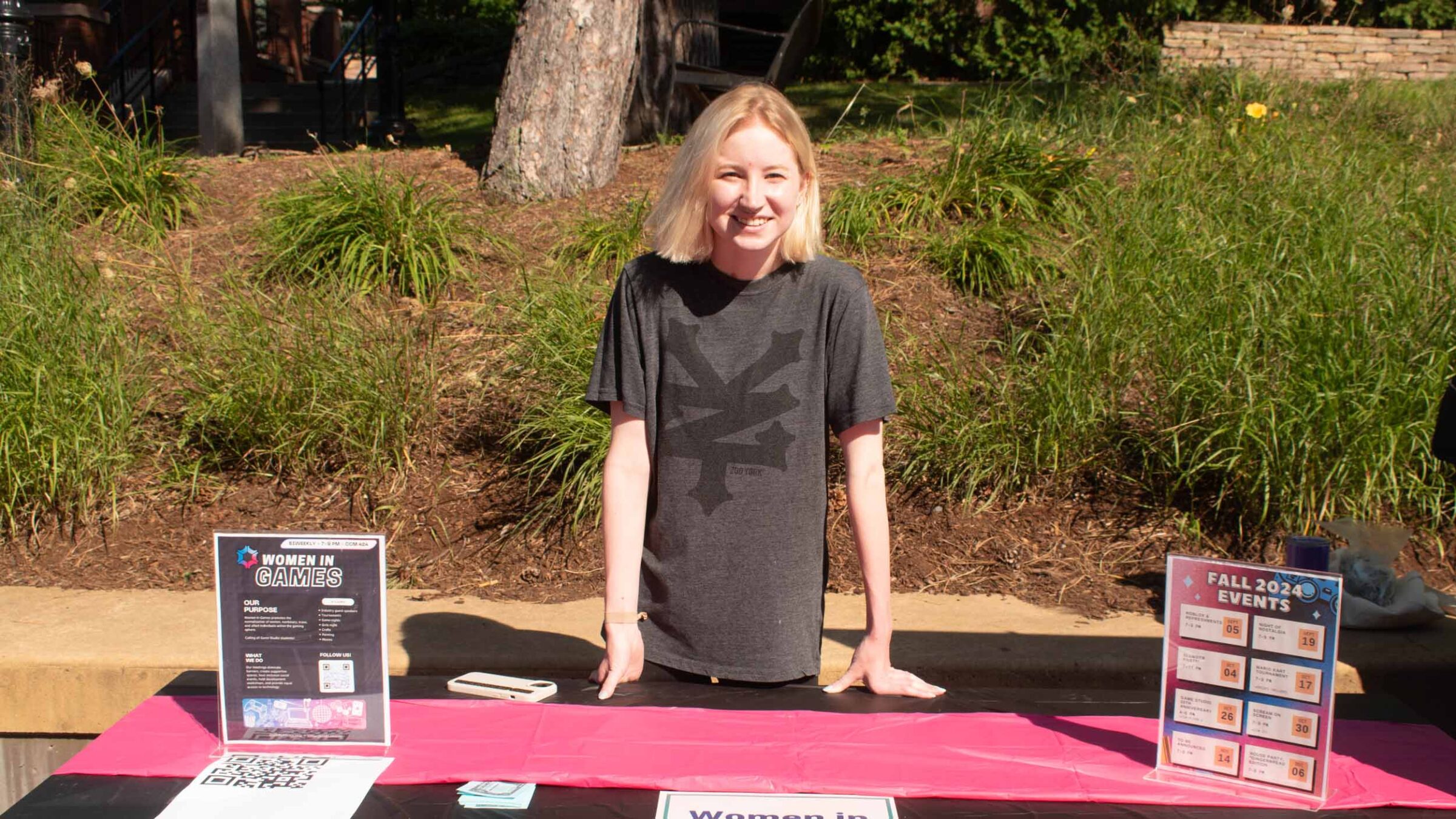 student standing behind the women in games club table at the activity fair outside