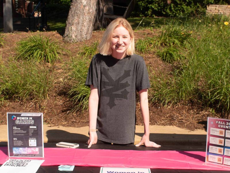 student standing behind the women in games club table at the activity fair outside