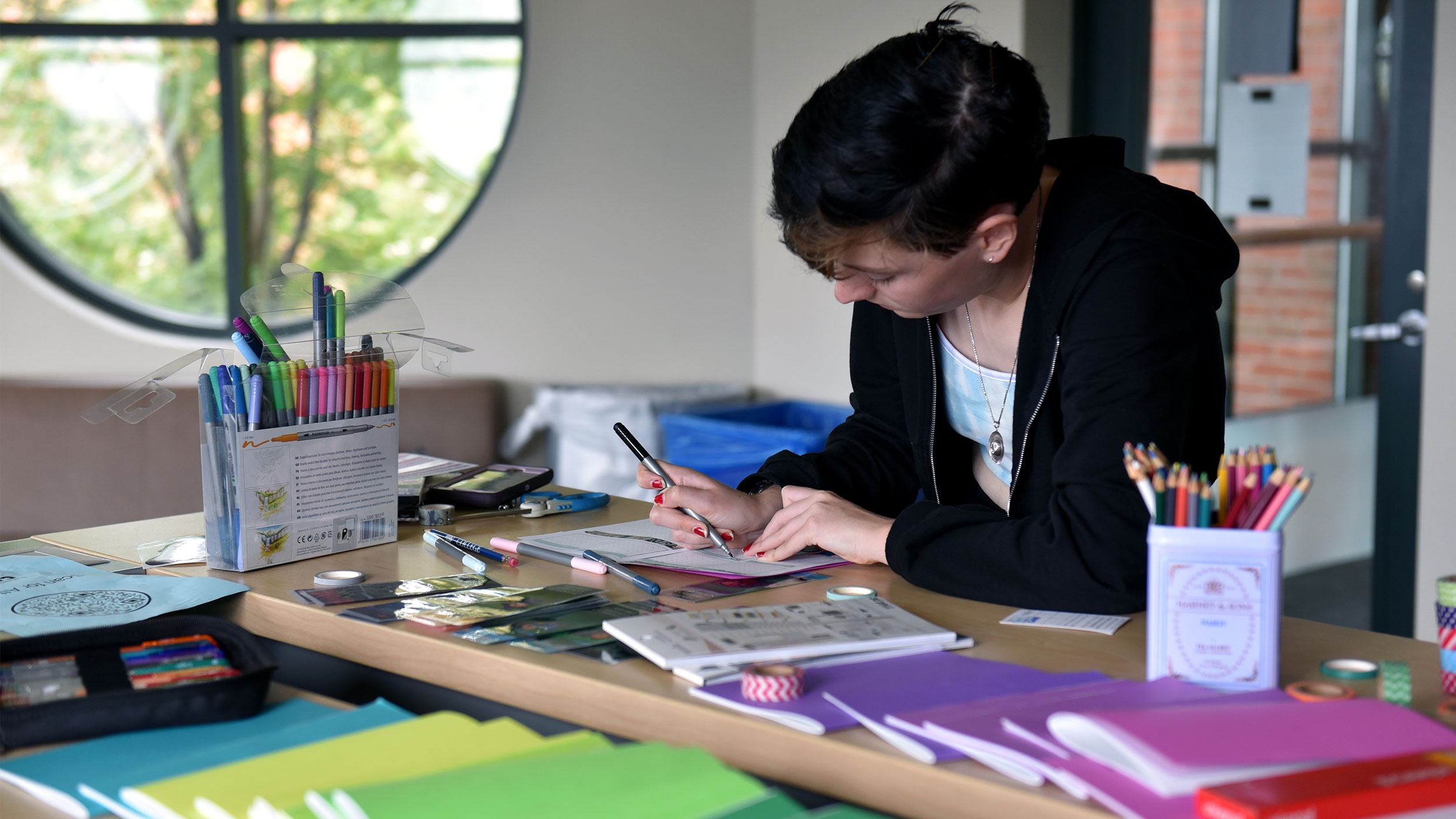 a student concentrates on writing in their gratitude journal. the table they're working on is covered in bright colored gratitude journals.