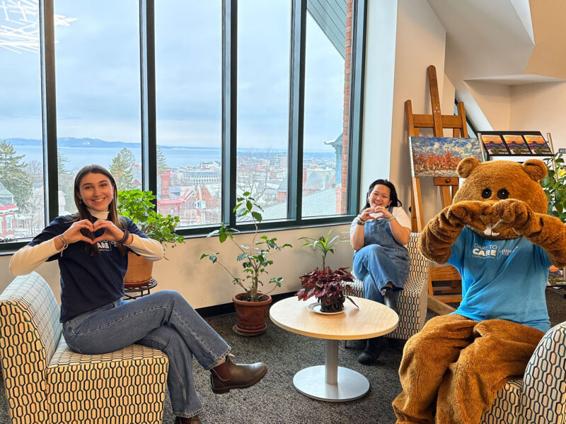 two students and chauncey the beaver mascot sit on chairs, making heart shapes with their hands. Lake Champlain is visible through the large windows behind them.