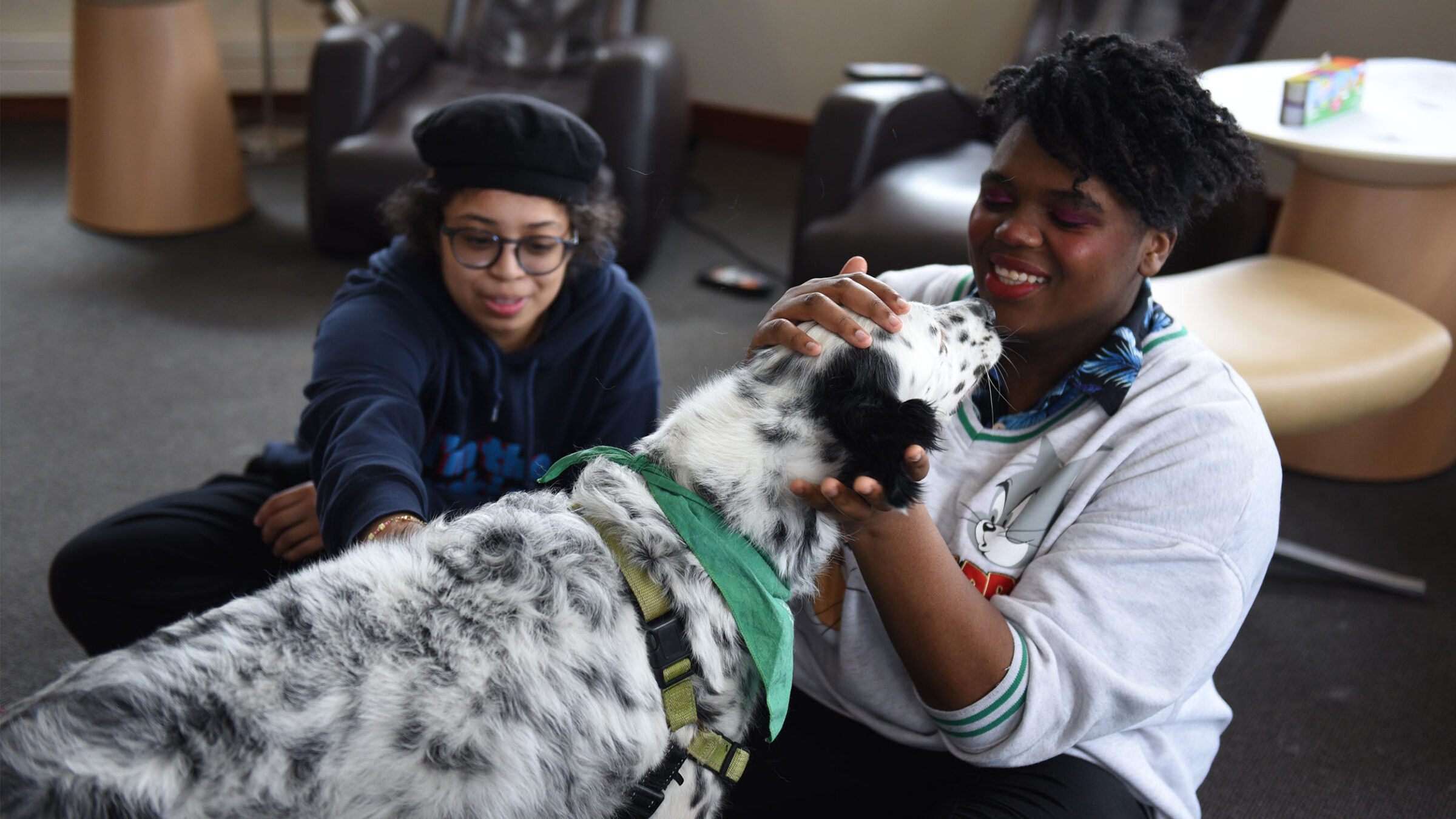 two students smile while petting a black and white spotted therapy dog in the SMART Space