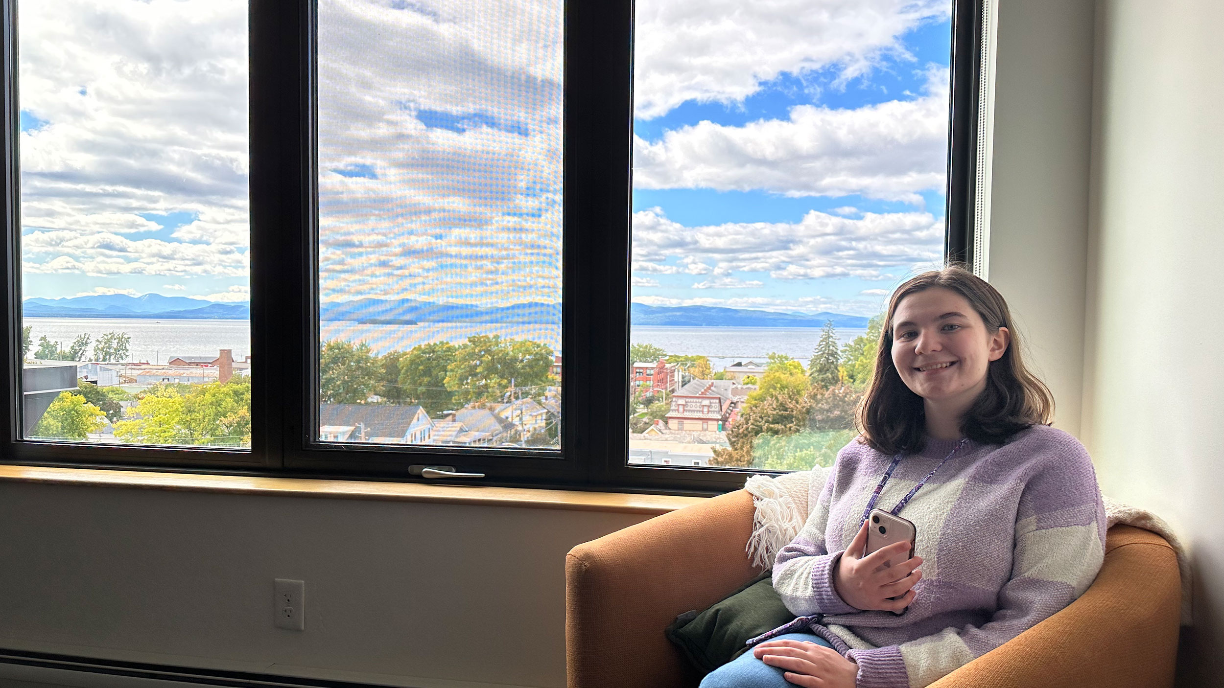 a student sits in a lounge chair holding their phone and smiling at the camera, with a panoramic view of lake champlain spanning through the window