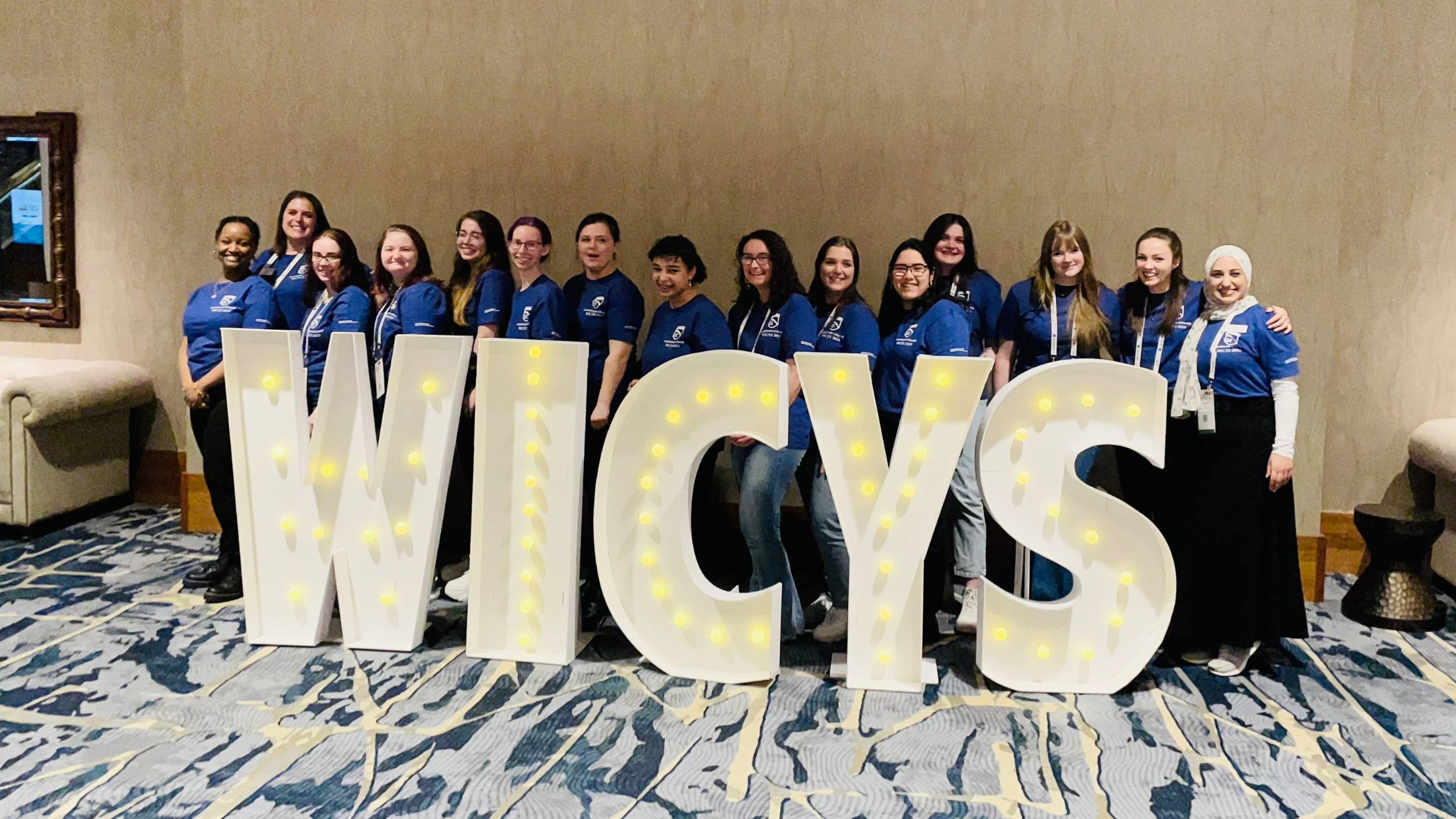 Female-identifying students from Champlain College pose with a light-up sign that reads "WICSY."