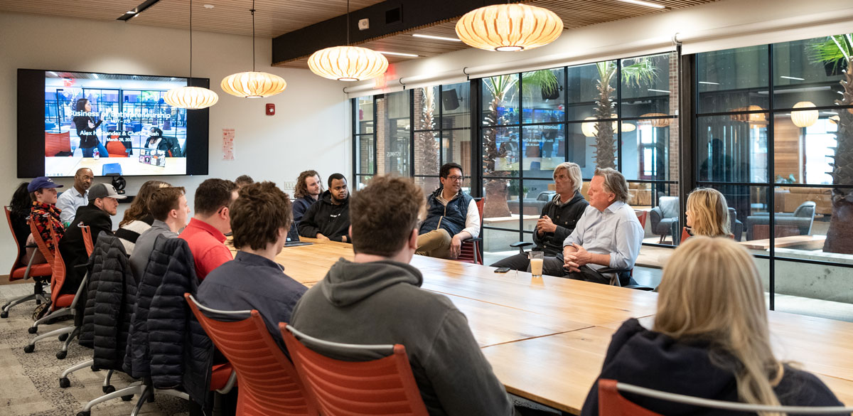 a group of students, staff, and faculty sitting in a board room having a discussion; a large tv screen shows a presentation