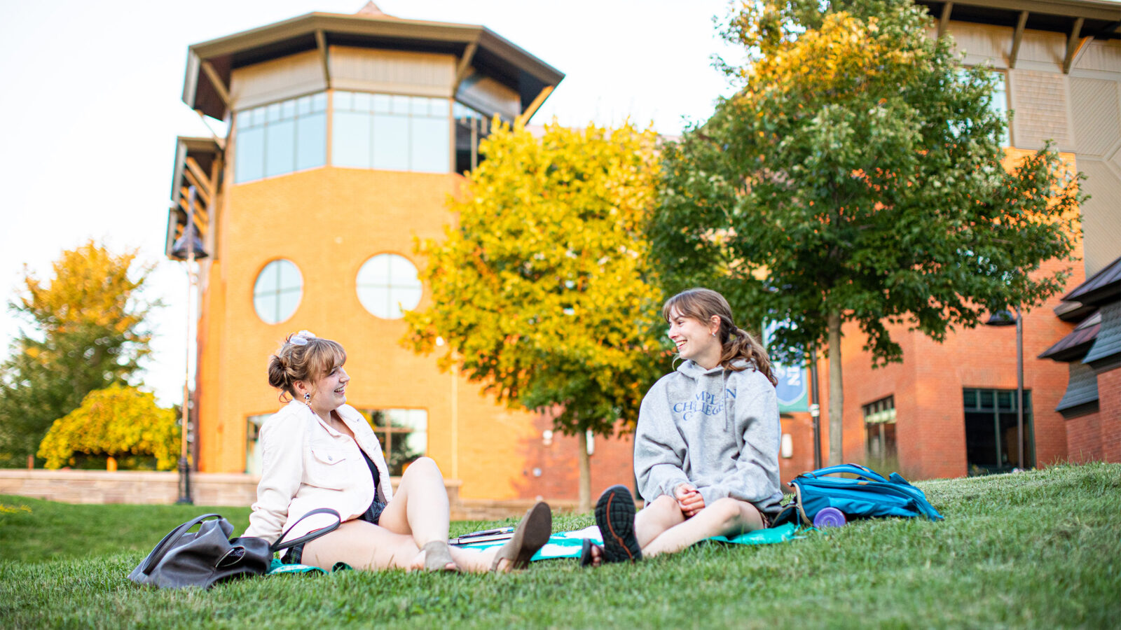 Two students sit on a blanket on Aiken Lawn outside the library. They are talking and relaxing on a sunny day.