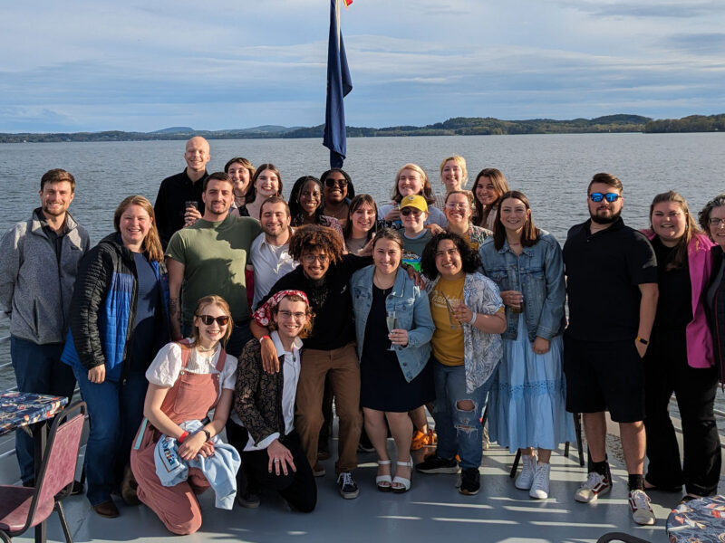 twenty students pose for a photo on the deck of the ethan allen boat cruise with lake champlain visible in the background