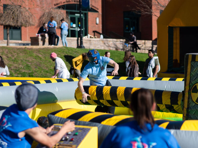 a student jumps over a big moving lever on a big yellow and black inflatable carnival obstacle course on the aiken lawn