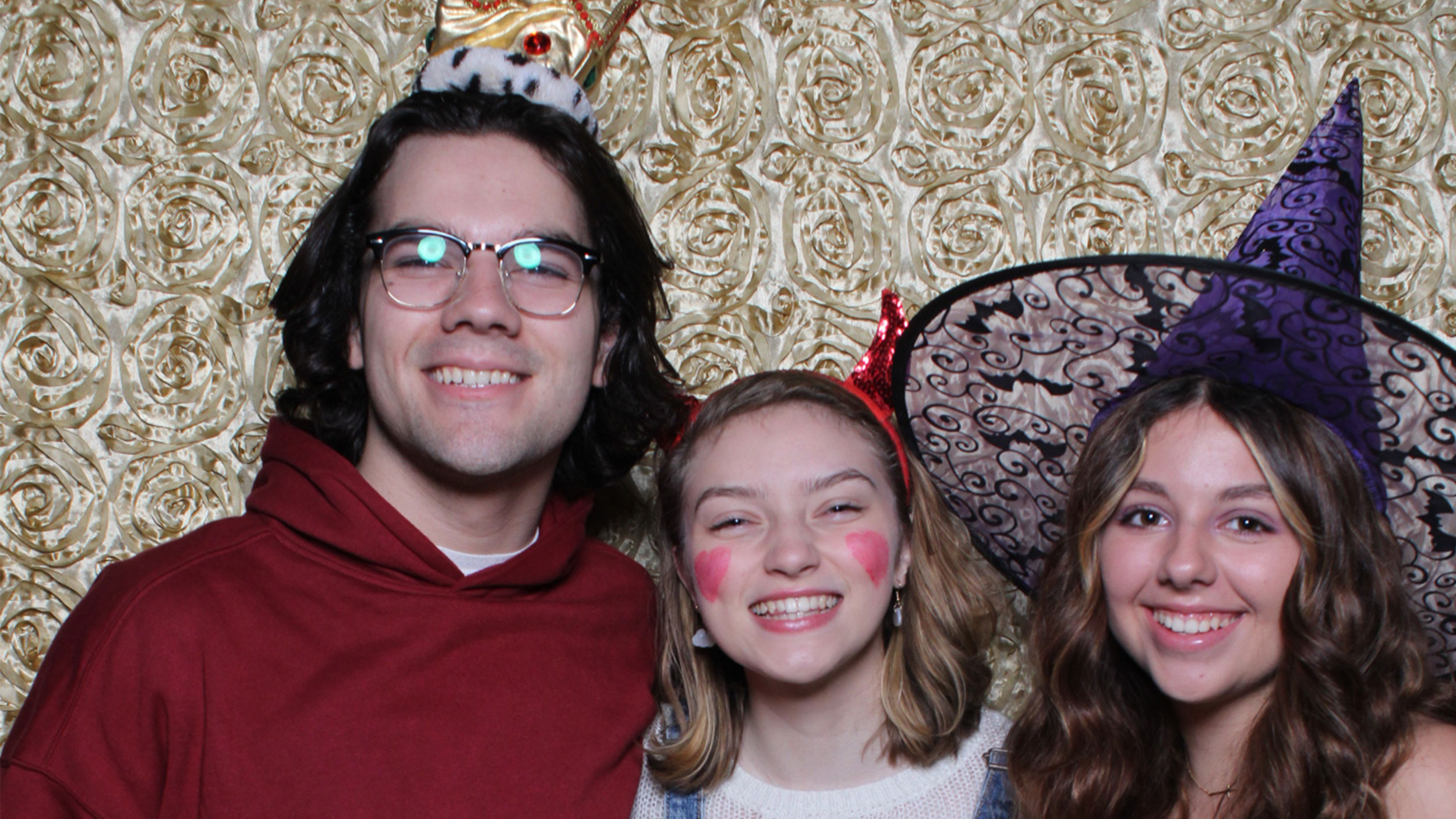 three students smile in their halloween costumes. one is wearing devil ears and one is wearing a witch hat