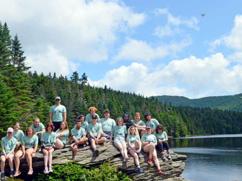a group of about 20 students sit on a huge rock in front of a big pond with green mountains and blue sky in the background