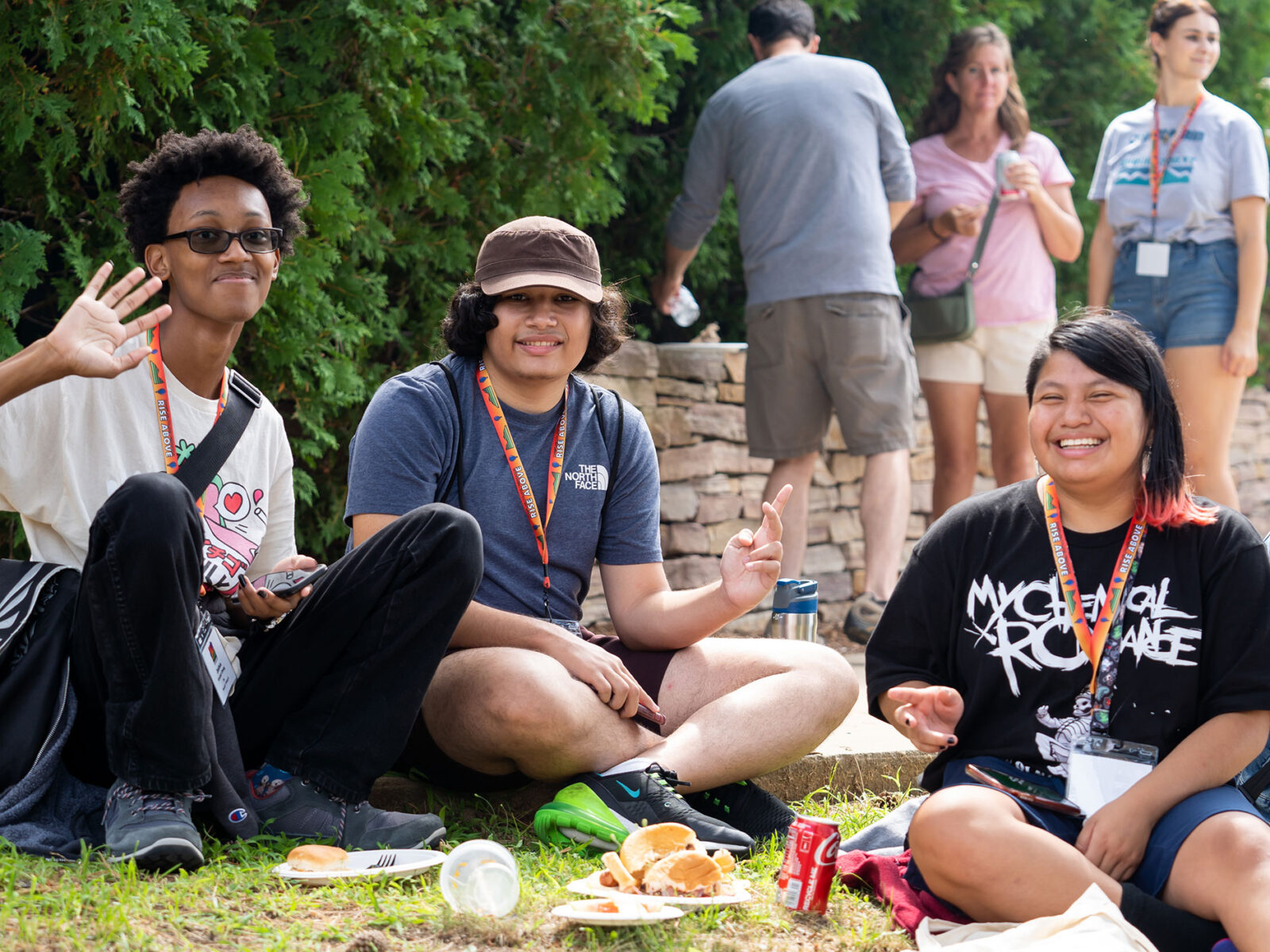 three students sit on the grass eating a BBQ lunch at orientation, waving and giving peace signs at the camera