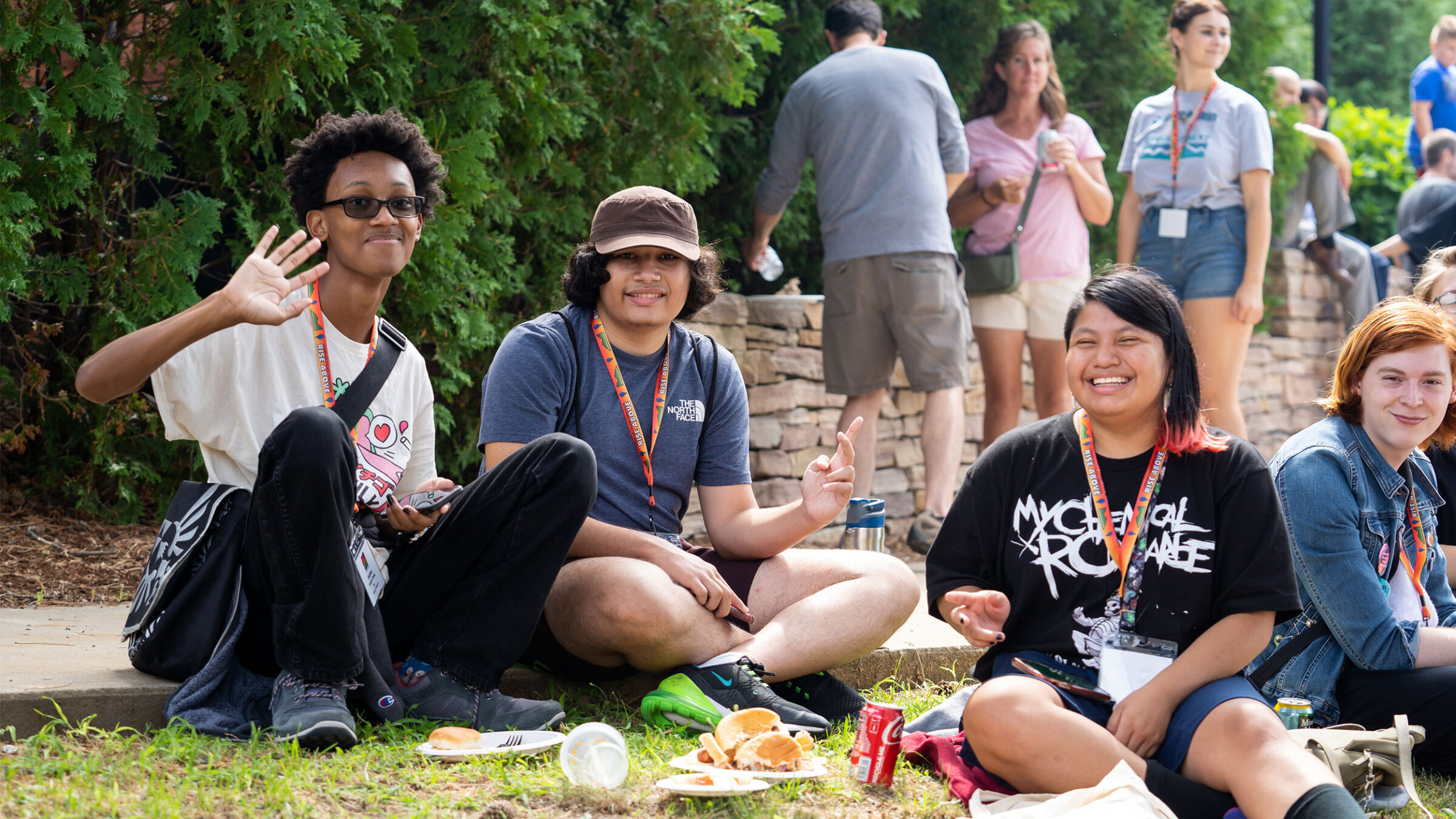 three students sit on the grass eating a BBQ lunch at orientation, waving and giving peace signs at the camera