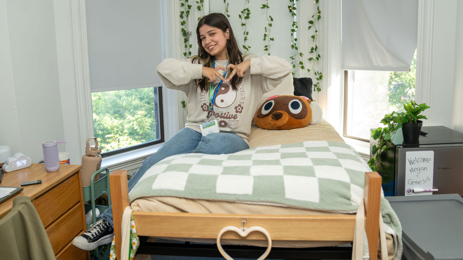 student makes a heart shape with their hands while sitting on their twin bed in a residence hall