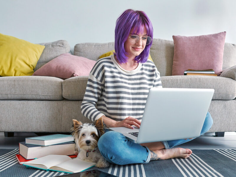 young woman with purple hair and a pet dog sit on the floor viewing a laptop