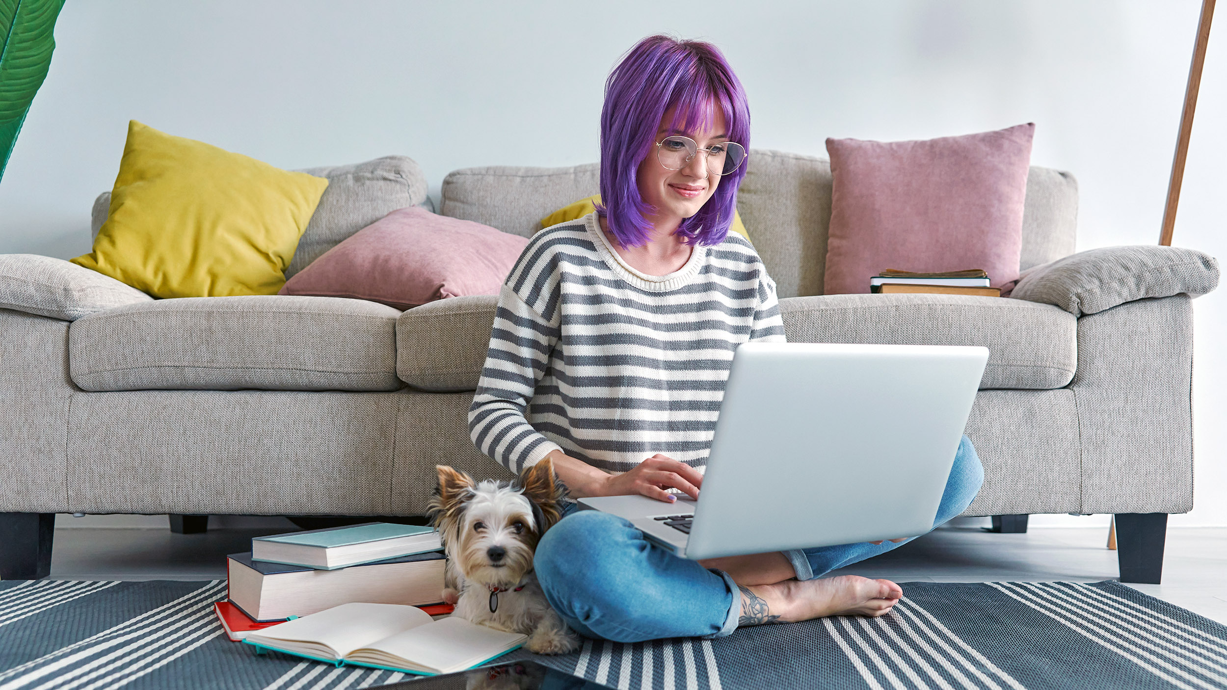 young woman with purple hair and a pet dog sit on the floor viewing a laptop