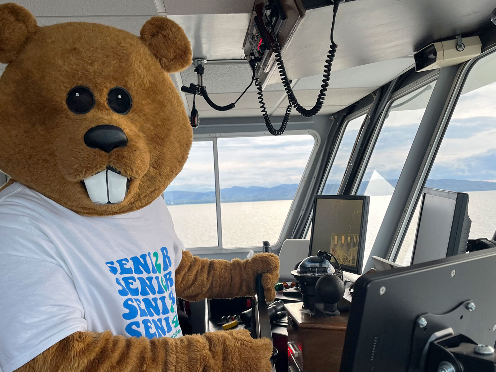 mascot chauncey t. beaver captains the ethan allen boat cruise with a lake and mountain view through the boat window