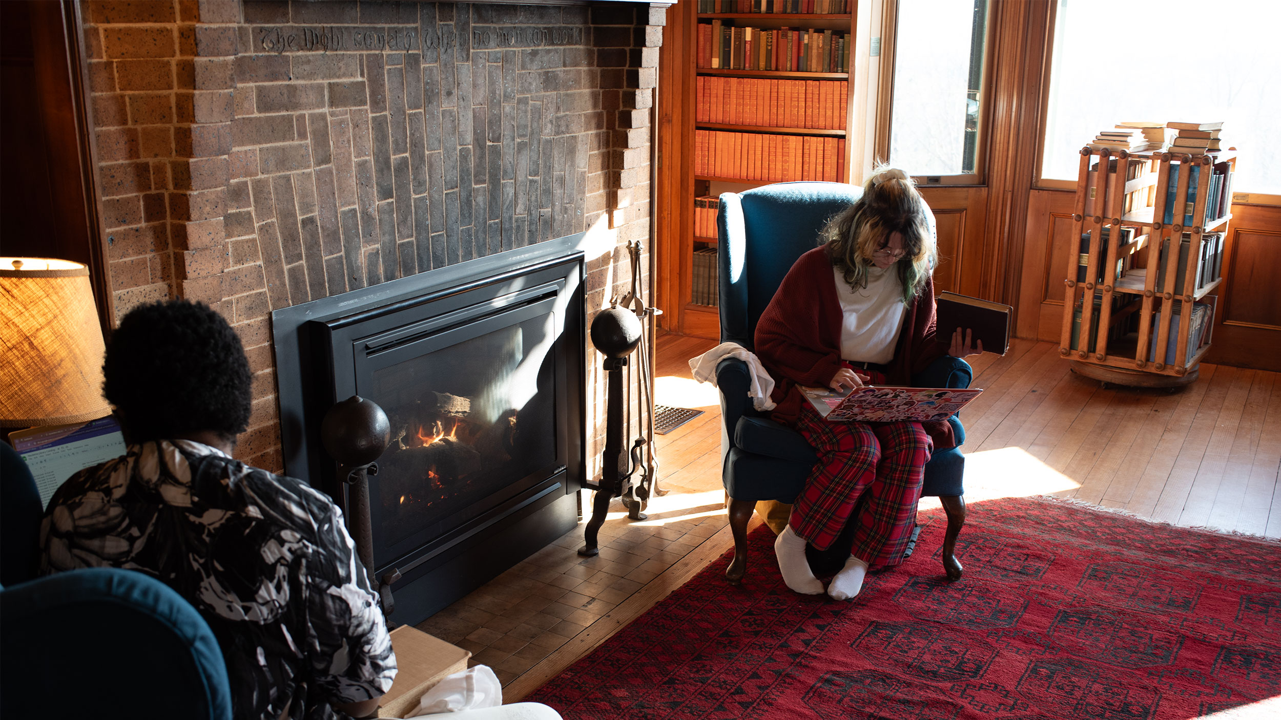 two students sit in chairs in front of a brick-surrounded fireplace in the library of an old estate