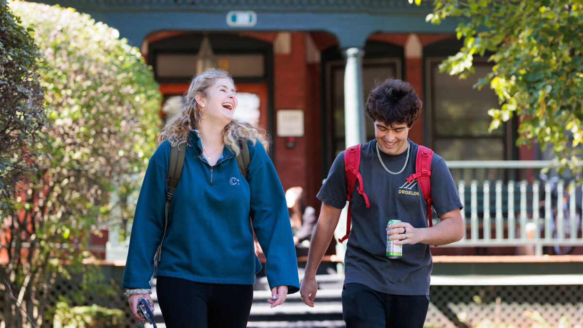 two students walk to class with their backpacks on, laughing, with greenery in the background